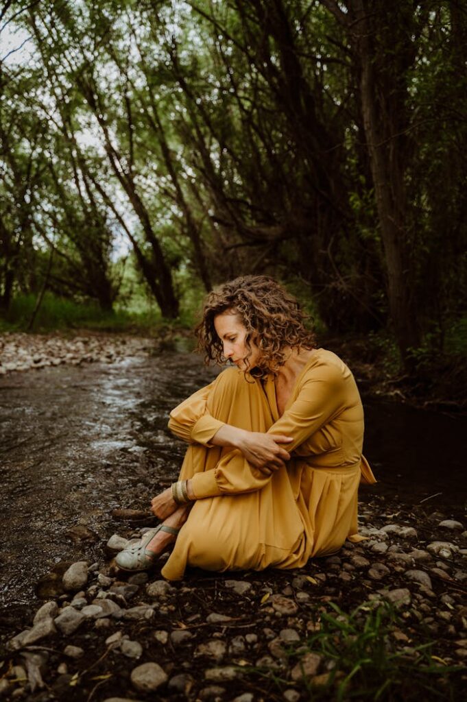 A woman in yellow sits pensively by a forest stream, reflecting amidst nature.